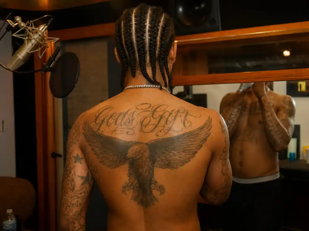 Post Malone tattoos and their meanings back piece — musician with braids from behind showing eagle wing shoulder tattoo in recording studio mirror