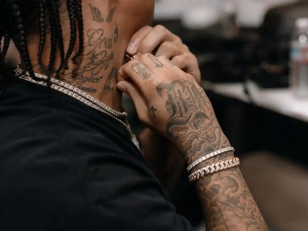 Post Malone tattoos and their meanings face ink — tattooed hands and neck script tattoos of musician with braids in green room, side angle no identifiable face