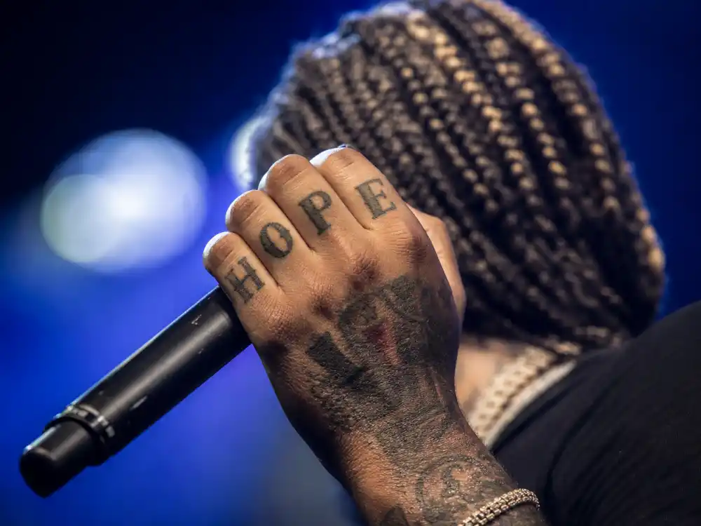 Post Malone tattoos and their meanings hand ink — close-up of tattooed fingers holding microphone with knuckle letter tattoos and braids visible from behind