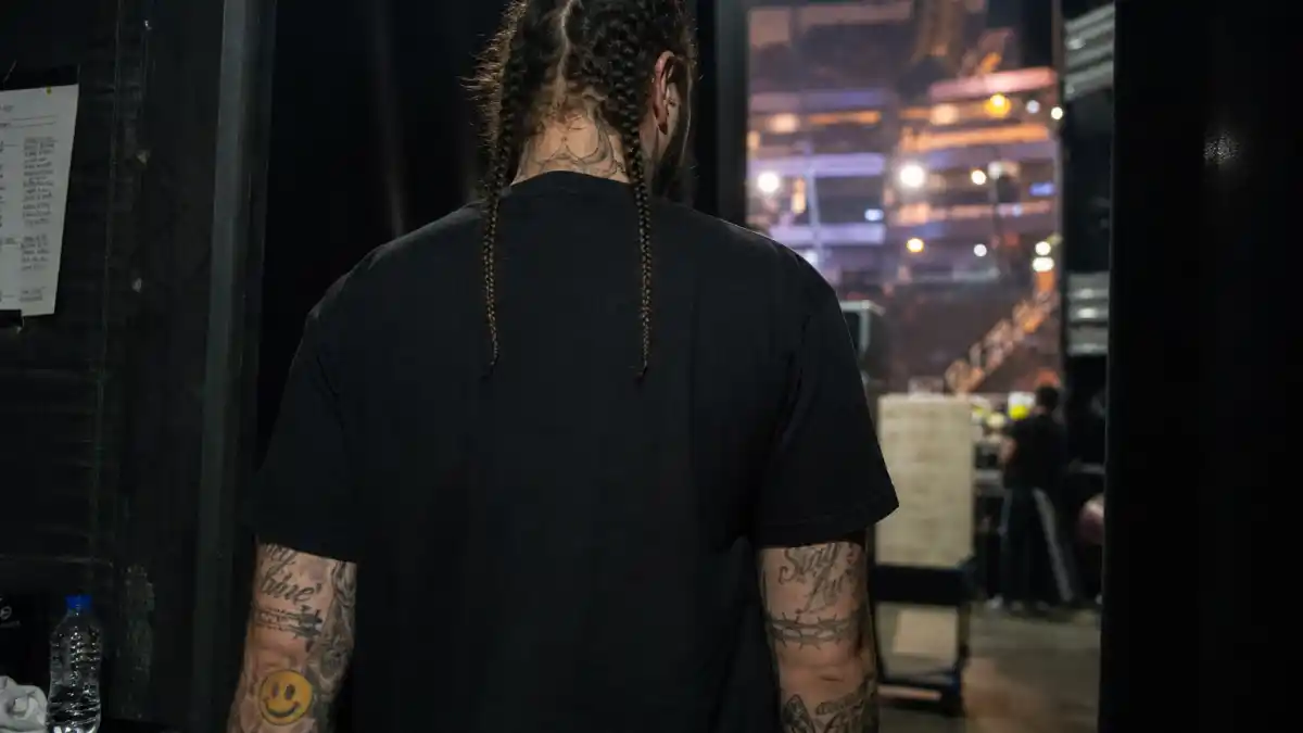 Post Malone tattoos and their meanings — musician with braided hair from behind showing barbed wire forearm tattoos and neck ink backstage at arena