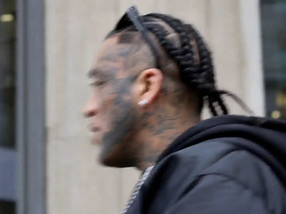 Post Malone tattoos and their meanings neck tattoos — motion-blurred side profile of braided musician showing heavy facial and neck script ink outdoors