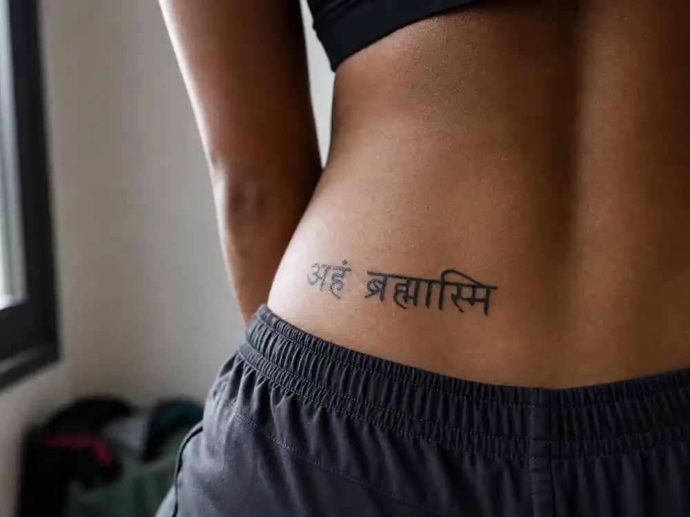 Rihanna tattoos and their meanings — Sanskrit script tattoo along hip bone photographed from behind in natural window light