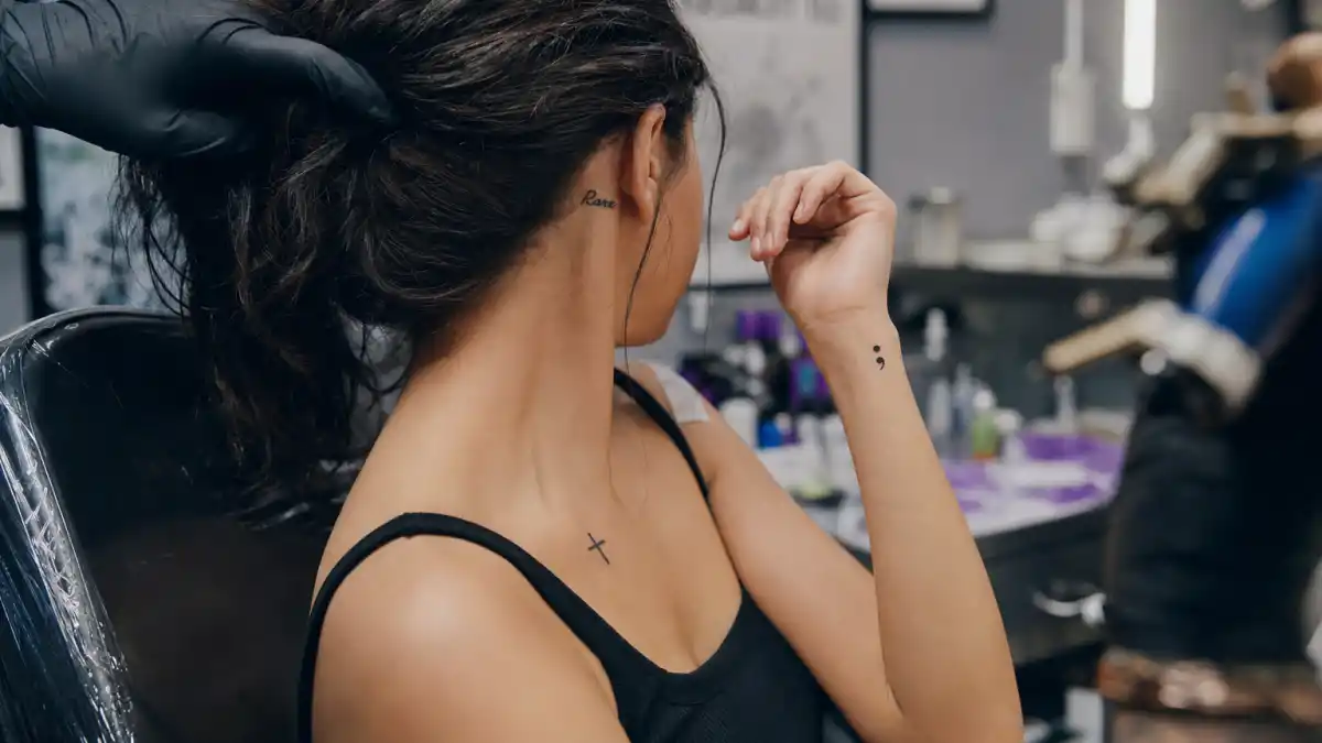 Selena Gomez tattoos and their meanings — pop star seen from behind in tattoo studio with Rare neck script, cross collarbone ink, and semicolon wrist visible