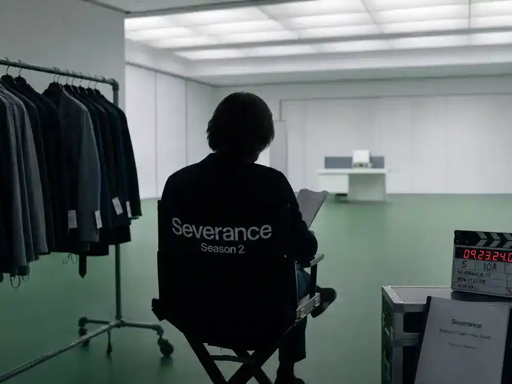 Severance season 2 cast and plot guide Adam Scott Mark Scout — actor silhouette from behind on director chair reviewing script at sterile corporate soundstage