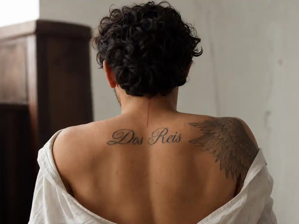 Shawn Mendes tattoos and their meanings — Dos Reis family surname back tattoo angel wing shoulder and red neck line tribute ink