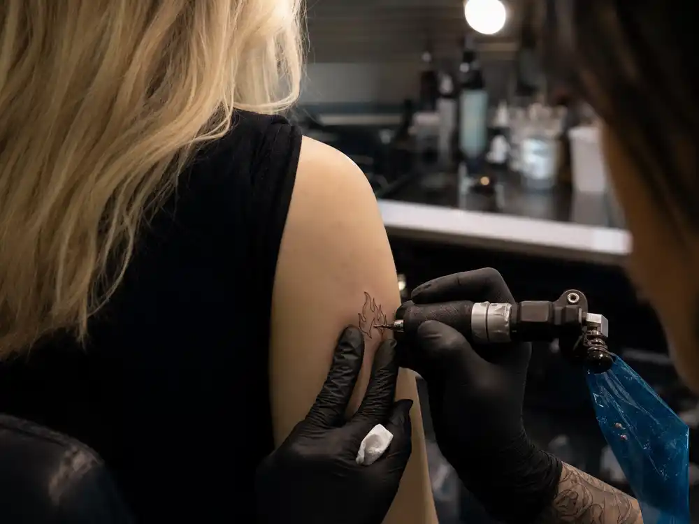 Sophie Turner tattoos and their meanings studio session — tattoo artist applying fine-line flame symbol tattoo during professional session