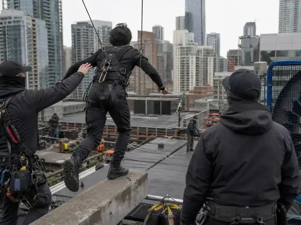 Spider-Man Brand New Day cast and characters guide Peter Parker — wire-rig stunt rehearsal on urban rooftop film set from behind, production crew visible