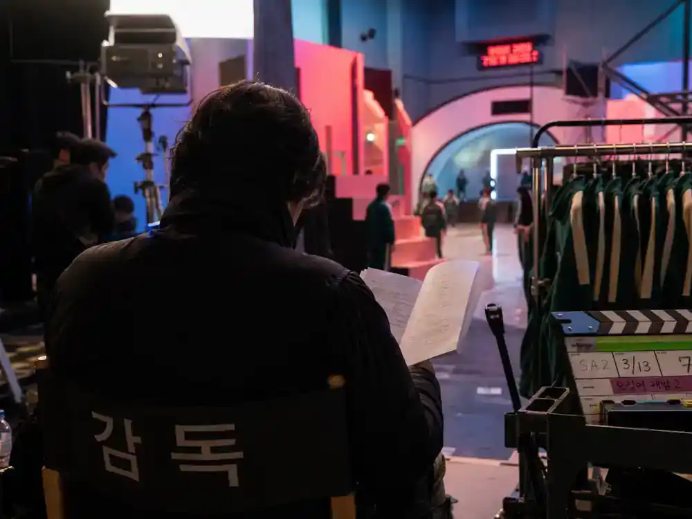 Squid Game cast and characters explained Gi-hun — lead actor silhouette from behind on director chair reviewing script on colorful TV game set soundstage