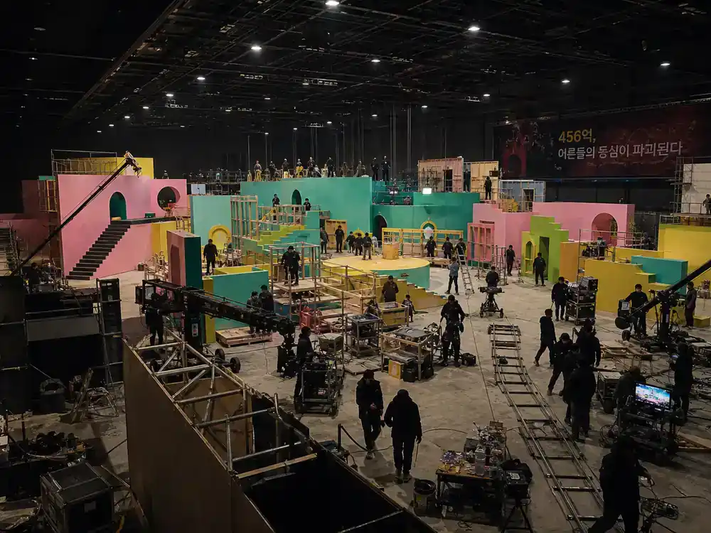 Squid Game season 2 behind the scenes facts Daejeon studio — massive indoor film warehouse with colorful game set construction, scaffolding, and crew silhouettes working under cranes