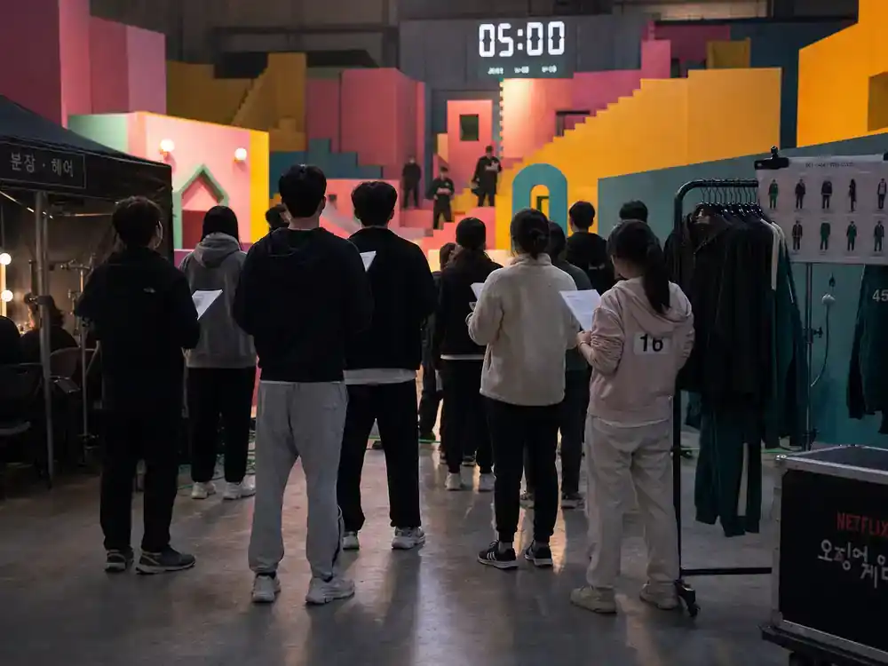 Squid Game season 2 behind the scenes facts ensemble cast — TV drama cast silhouettes from behind rehearsing on colorful geometric game set with wardrobe racks and makeup tents