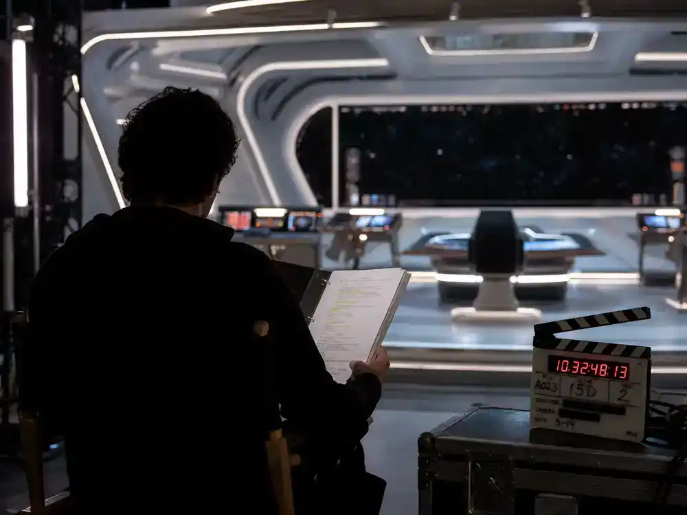 Star Trek universe 2026 shows and cast guide Strange New Worlds — actor silhouette from behind on director chair reviewing script on starship bridge TV soundstage