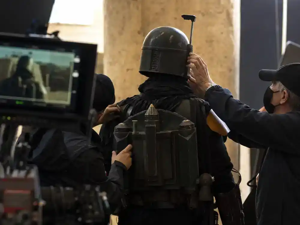 Star Wars upcoming movies and cast guide Mandalorian and Grogu — actor silhouette from behind in armored bounty hunter costume on film set with crew adjusting helmet