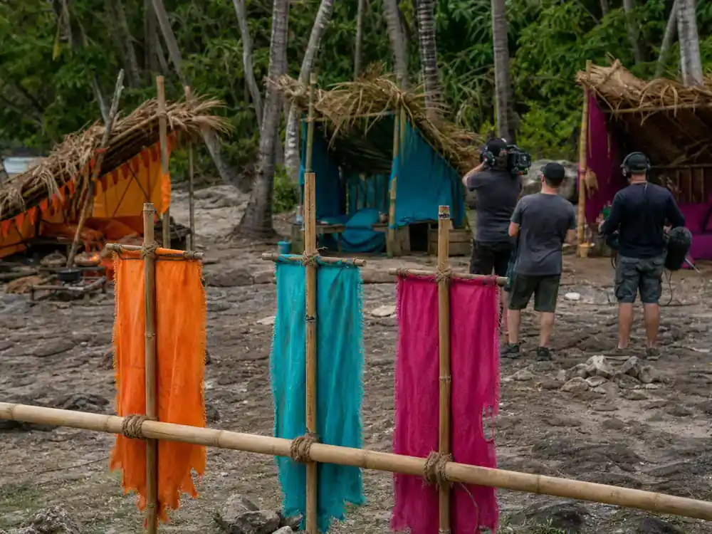 Survivor 50 cast and season preview guide tribes — orange teal and magenta tribe buffs at three bamboo camp shelters on tropical island