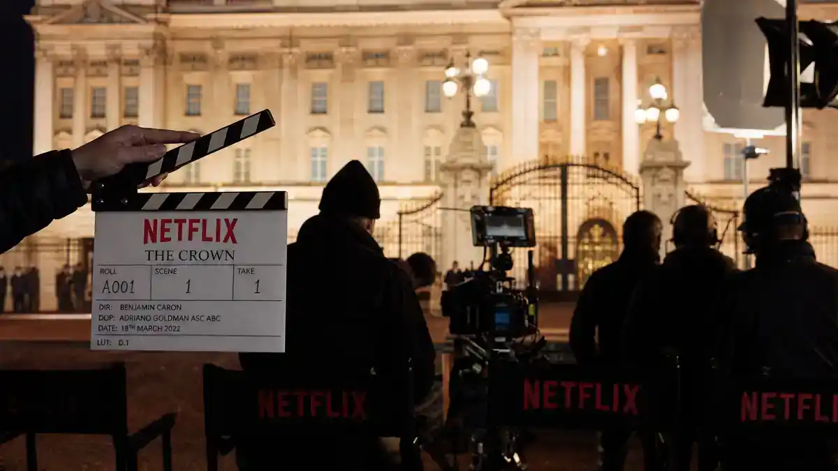 The Crown cast vs real royals guide — Netflix royal drama production set with clapperboard director chairs and palace facade backdrop, editorial TV reference