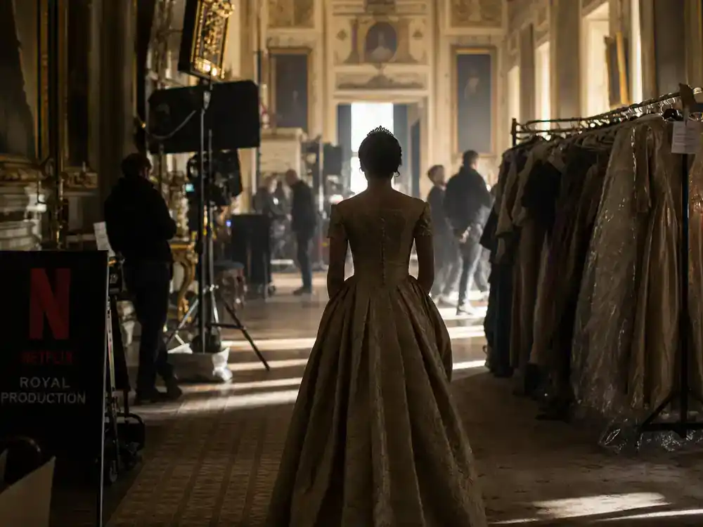 The Crown cast vs real royals guide Queen Elizabeth cast — actress silhouette from behind on palace corridor period drama film set with wardrobe racks