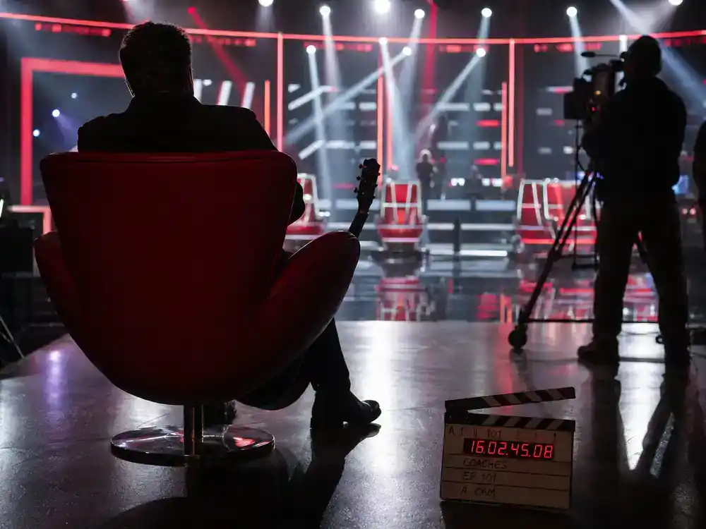 The Voice 2026 coaches and cast guide Adam Levine — musician silhouette from behind in red coach chair on singing competition soundstage with clapperboard