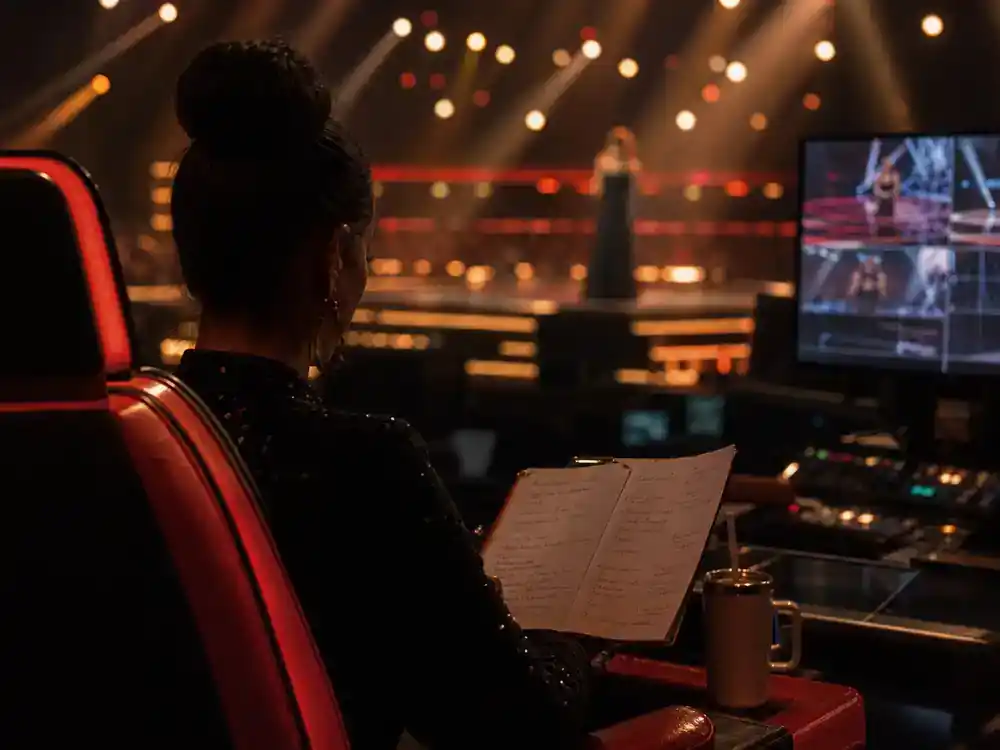 The Voice 2026 coaches and cast guide Kelly Clarkson — singer silhouette from behind in red coach chair reviewing notes on TV studio set