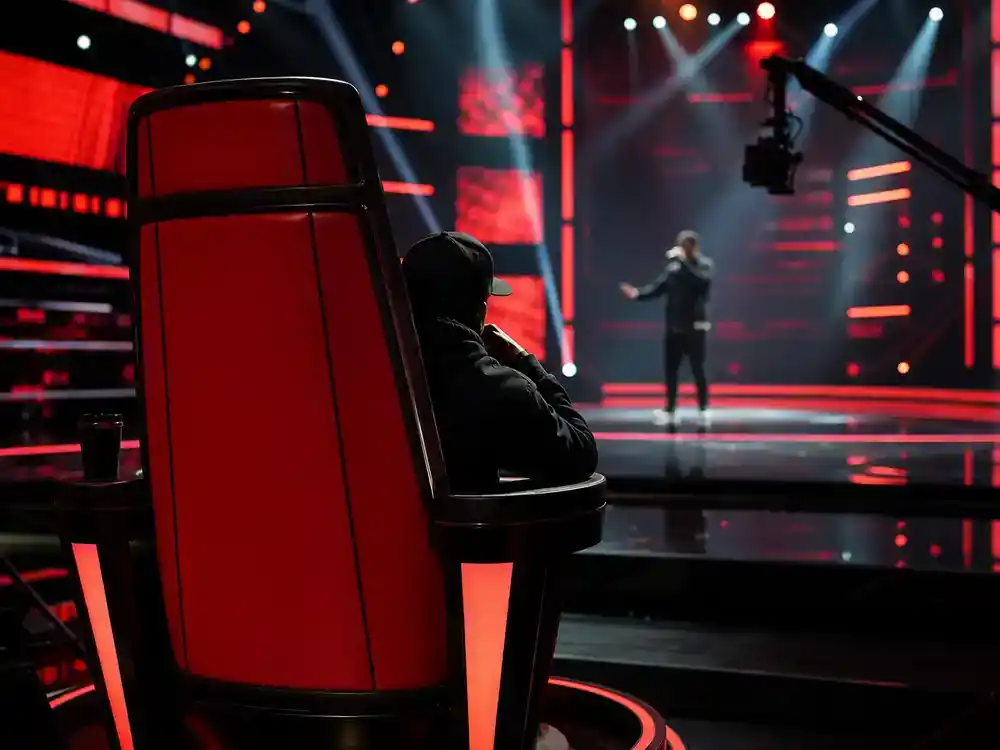 The Voice 2026 coaches and cast guide Queen Latifah — performer silhouette from behind in red coach chair on TV studio set with production monitors