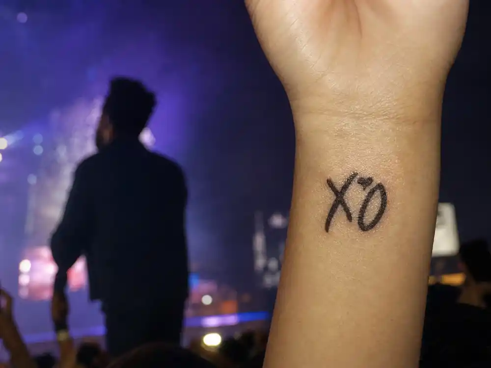 The Weeknd tattoos and their meanings — close-up of fan forearm XO script tattoo at concert with singer silhouette on stage