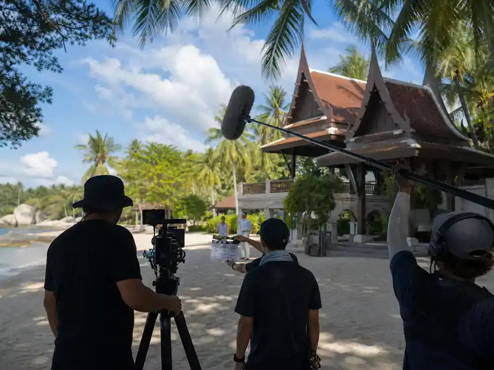 The White Lotus filming locations season guide Thailand — TV production crew filming on lush tropical resort beach with palm trees camera boom and clapperboard