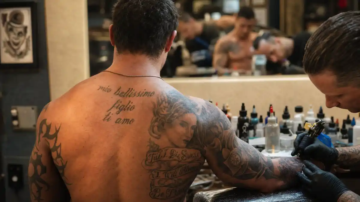Tom Hardy tattoos and their meanings — British actor seen from behind in tattoo studio with extensive arm and back ink visible