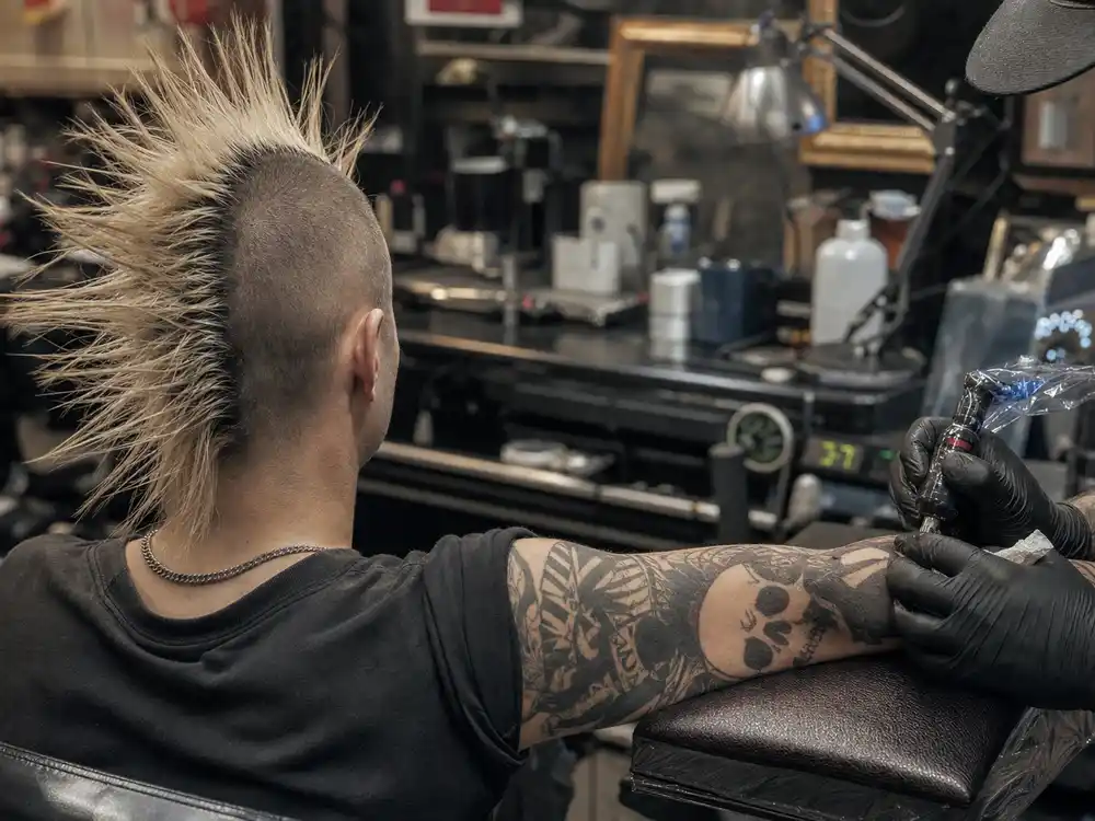 Travis Barker tattoo collection guide arm sleeve — close-up of black grey skull and script forearm tattoos on punk drummer in studio mirror
