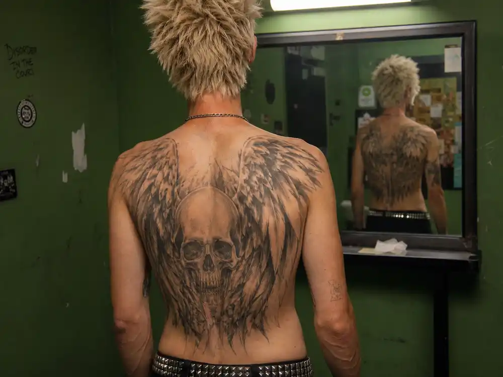 Travis Barker tattoo collection guide back piece — mohawk drummer from behind showing large skull and wing shoulder blade tattoos in green room
