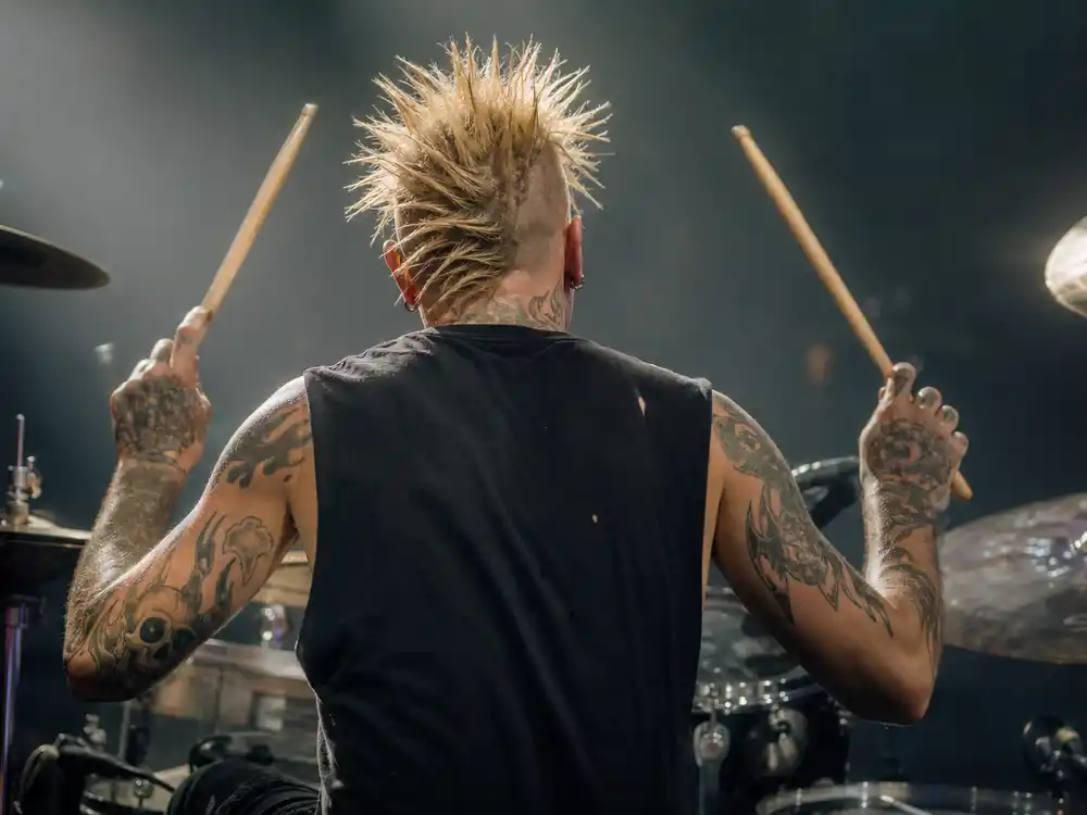 Travis Barker tattoo collection guide drum kit — shirtless punk drummer from behind at kit showing full back and arm tattoo coverage under stage lights