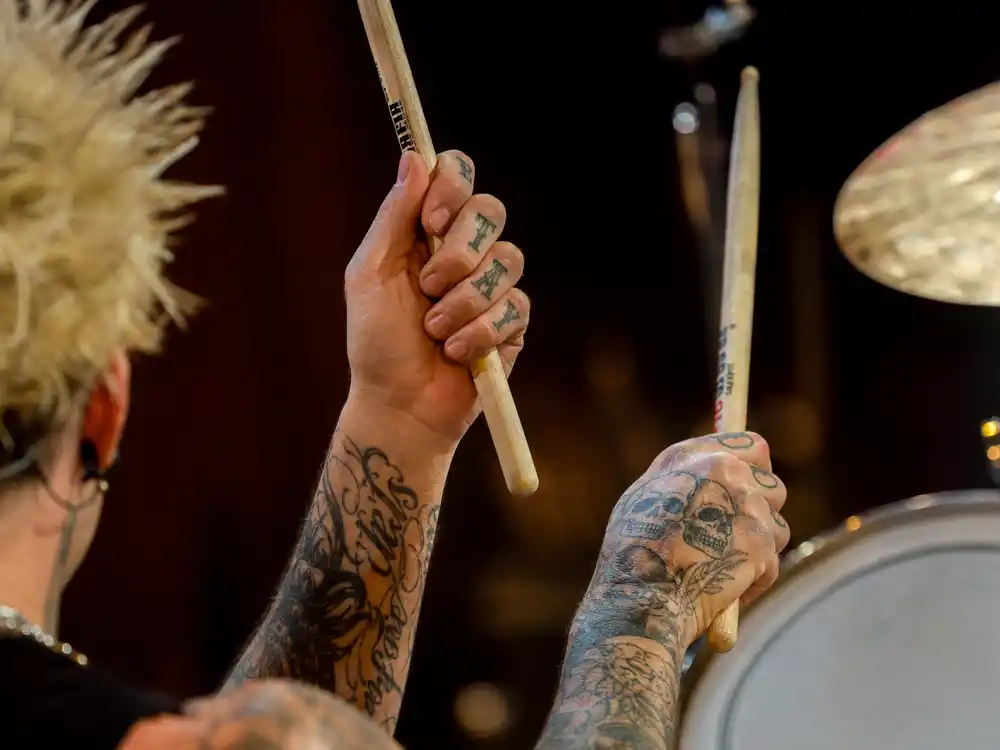 Travis Barker tattoo collection guide hand ink — close-up of tattooed knuckles and fingers holding drumsticks with letter script visible