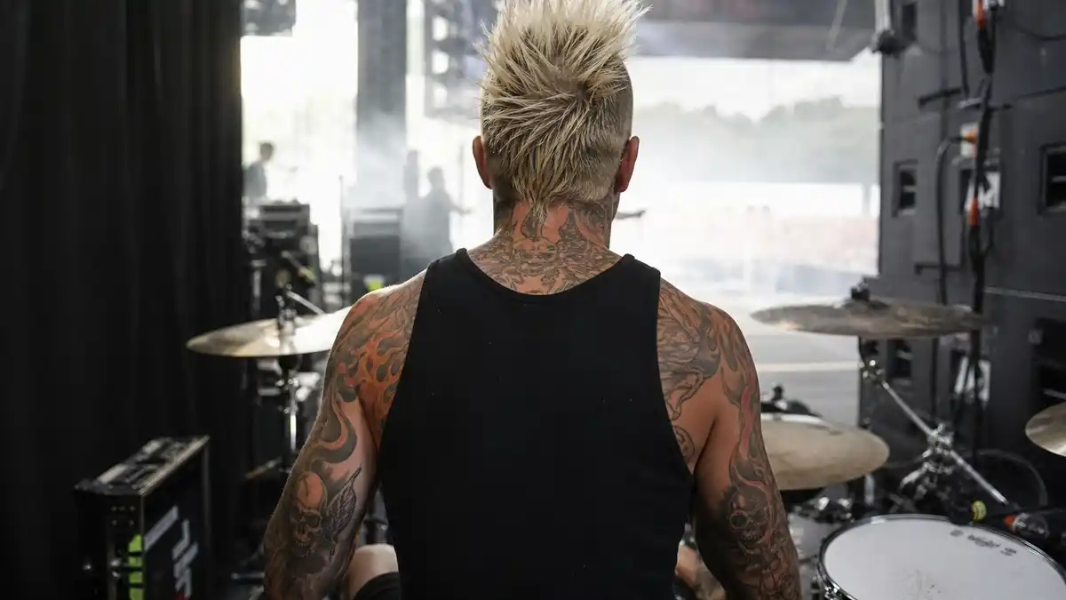 Travis Barker tattoo collection guide — punk drummer with blonde mohawk from behind at drum kit showing full arm sleeves and back ink on stage