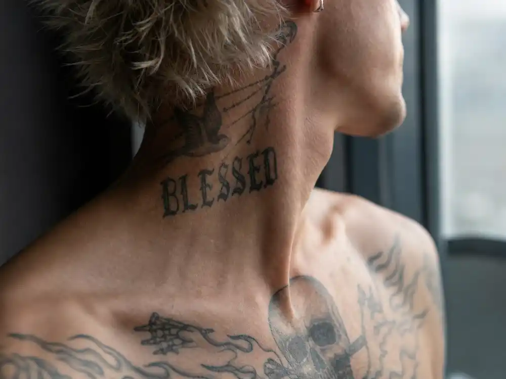 Travis Barker tattoo collection guide neck tattoo — side profile blurred mohawk drummer showing BLESSED script and cross neck ink outdoors