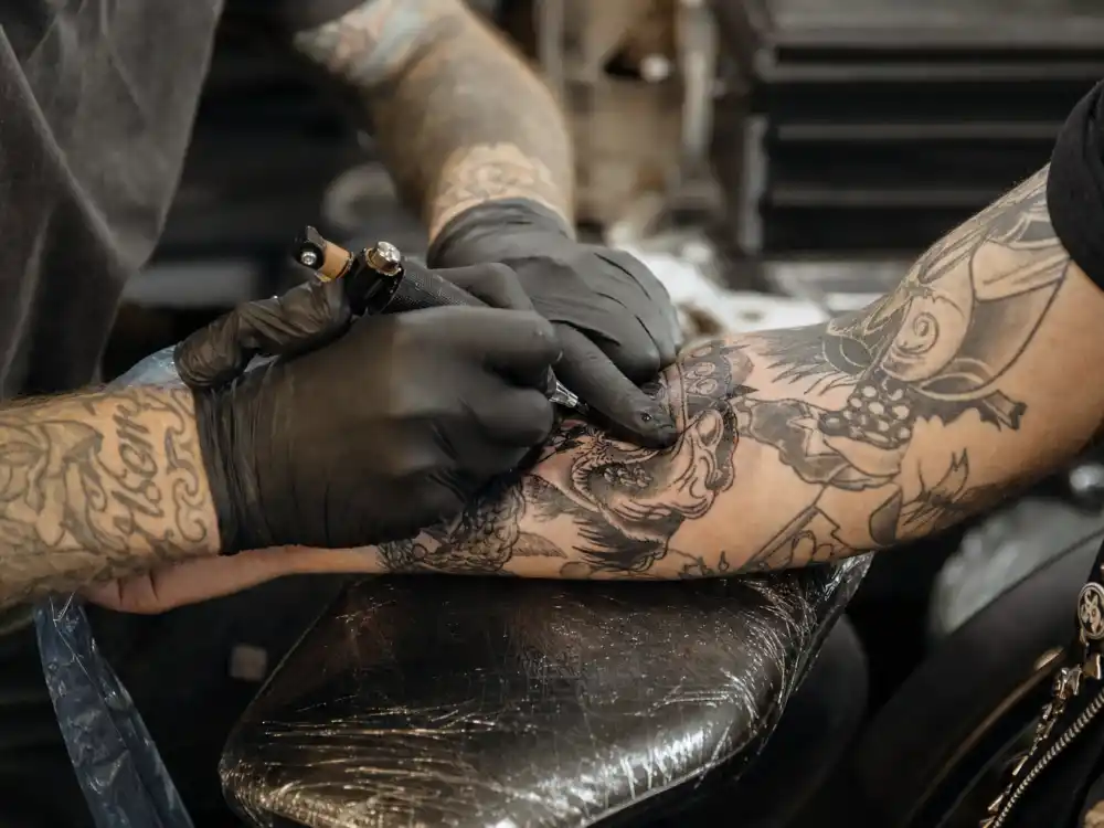 Travis Barker tattoos and their meanings — tattoo artist working on client forearm in professional studio session with existing sleeve ink