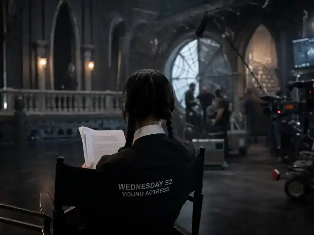 Wednesday season 2 cast and new characters guide Jenna Ortega — lead actress silhouette from behind on director chair reviewing script on gothic soundstage
