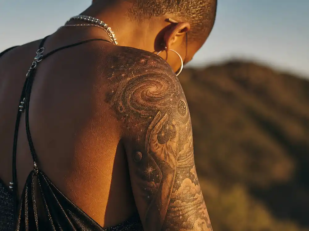 Willow Smith tattoos and their meanings — hand reaching into galaxy stars shoulder sleeve tattoo photographed from behind at golden hour