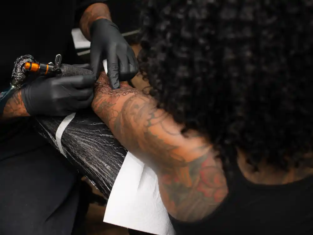 Wiz Khalifa tattoos and their meanings — tattoo artist working on client forearm in professional studio session with existing colorful sleeve ink