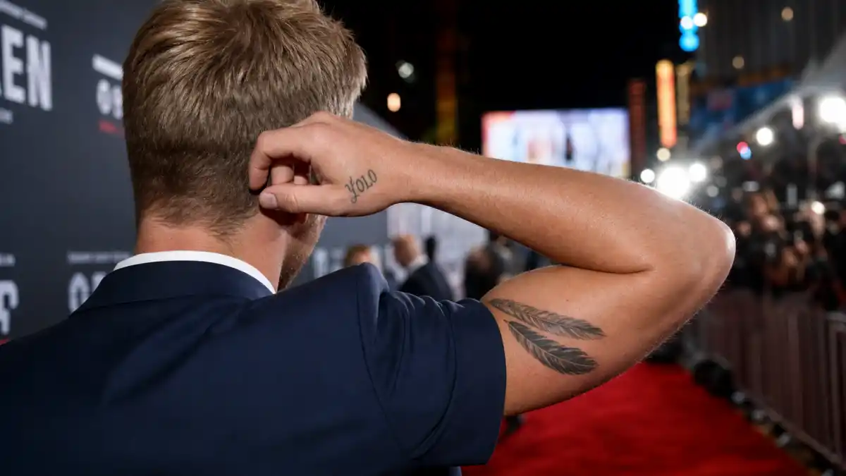 Zac Efron tattoos and their meanings — athletic blond actor from behind at premiere with YOLO hand tattoo and feather bicep ink visible
