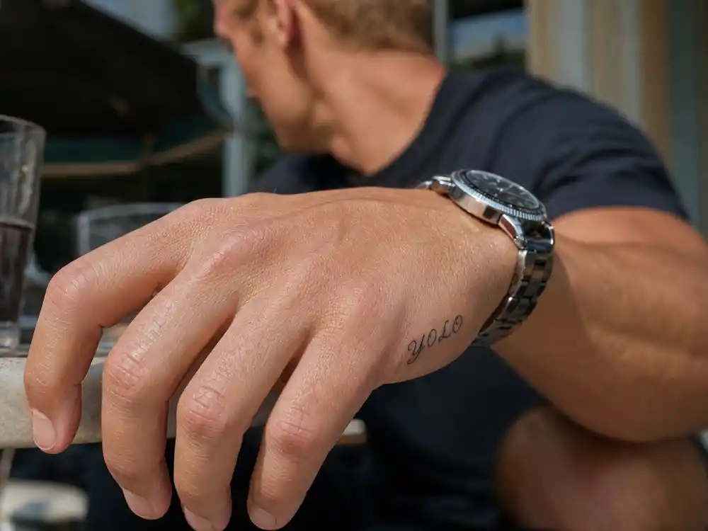 Zac Efron tattoos and their meanings — close-up of YOLO script tattoo on outer right hand near pinky finger at outdoor cafe