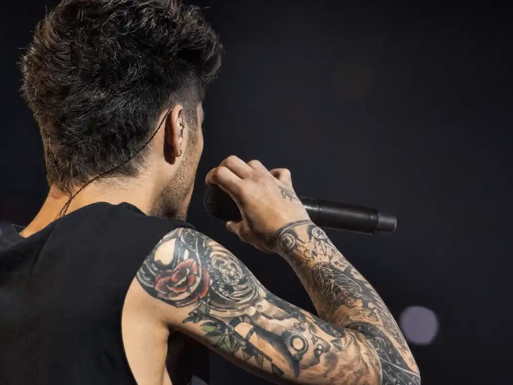 Zayn Malik tattoos and their meanings arm sleeve — red rose padlock and geometric blackwork forearm tattoos over-shoulder stage angle