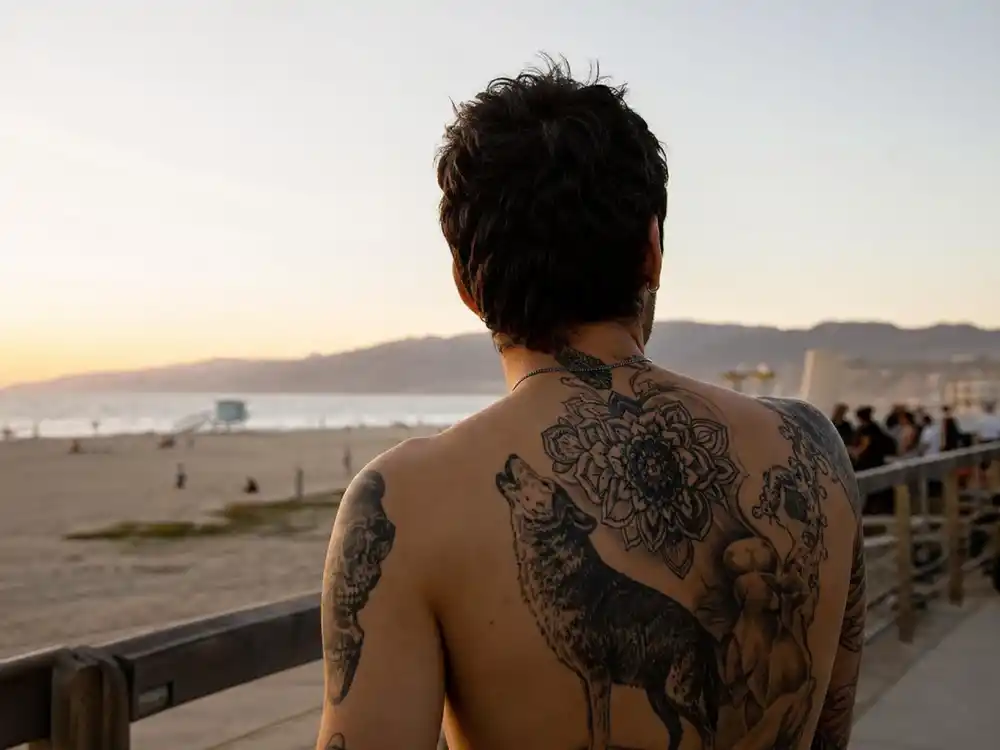 Zayn Malik tattoos and their meanings back tattoos — shirtless back view showing wolf silhouette and floral blackwork across shoulder blades at boardwalk