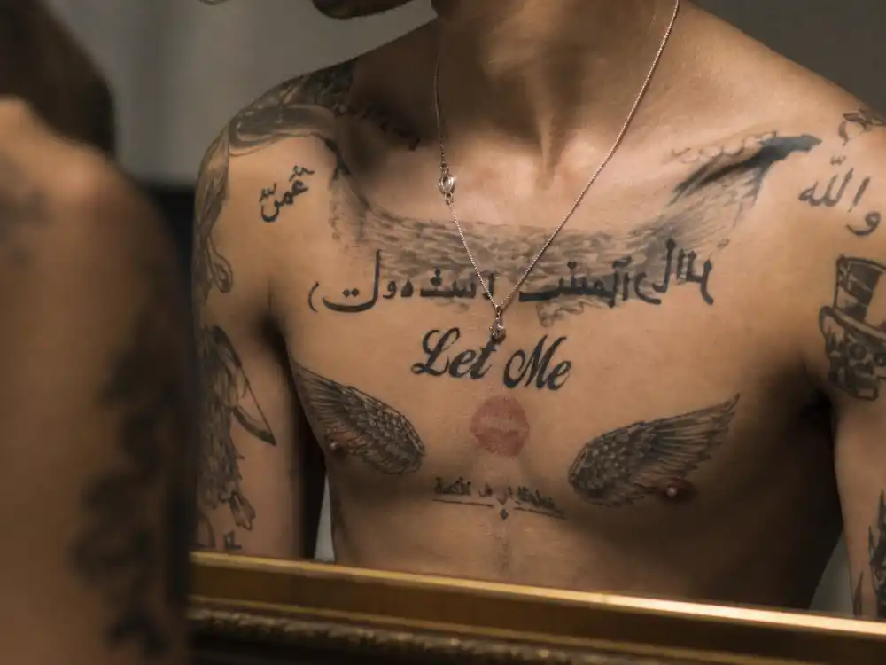 Zayn Malik tattoos and their meanings chest script — Arabic lettering and Let Me sternum tattoo mirror close-up on shirtless torso